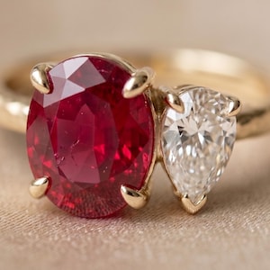 May include: A gold ring featuring a large oval-cut deep red ruby and a smaller pear-cut white diamond set side-by-side. The band has a textured finish and the stones are secured with gold prongs, creating a two-stone design on a yellow gold band.