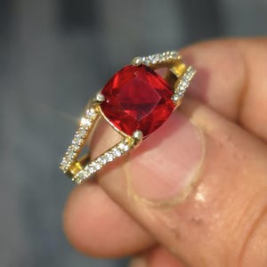 May include: A gold ring featuring a large, square-shaped red gemstone surrounded by smaller, clear gemstones.
