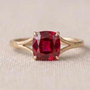 May include: A gold ring featuring a cushion-cut deep red gemstone set in a four-prong setting. The band has a split shank design that tapers as it connects to the central stone, creating a clean and balanced look for this piece of jewellery.