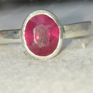 May include: A silver ring with a red gemstone set in a bezel setting. The ring is on a white surface.