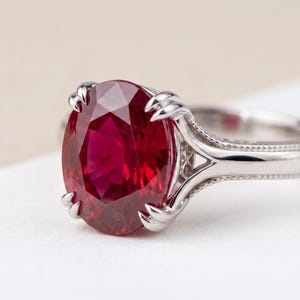 Op de afbeelding: Een zilverkleurige ring met een grote, ovaal geslepen, dieprode edelsteen die door vier pootjes wordt vastgehouden. De metalen band heeft een verfijnde korrelige textuur langs de randen, die leidt naar de centrale zetting die de rode steen vasthoudt.