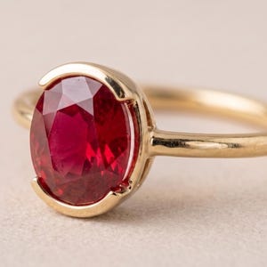 Op de afbeelding: Een gouden ring met een gefacetteerde, ovaal geslepen rode robijn in een gedeeltelijke zetting. De ringband is glad en gepolijst en heeft een warme geelgouden kleur die contrasteert met de diepe, levendige rode kleur van de centrale steen.