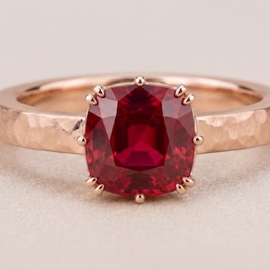 Op de afbeelding: Een roségouden ring met een dieprode edelsteen in kussenslijpsel, gezet in een klauwzetting. De ringband heeft een opvallende gehamerde textuur, waardoor een reeks kleine, reflecterende inkepingen over het oppervlak van de metalen band ontstaat.