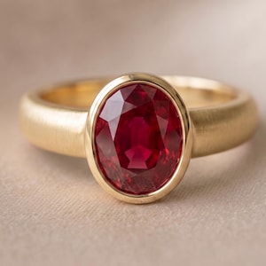 May include: A gold ring featuring a large, oval-cut, deep red ruby gemstone set in a smooth bezel setting. The band of the ring has a matte, brushed finish, providing a contrast to the polished gold bezel surrounding the faceted red stone.