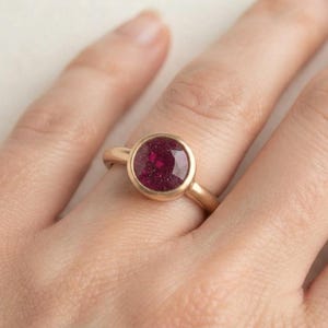 May include: A ring featuring a round, deep magenta-colored stone with a subtle glittery texture, set in a smooth, polished gold-toned metal band. The circular gemstone is held securely in a bezel setting, creating a clean and simple profile.
