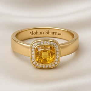 May include: A gold ring with a square, yellow gemstone at the center, surrounded by a halo of smaller, clear stones. The ring band is engraved with the name "Mohan Sharma". The ring is set against a soft, white background.