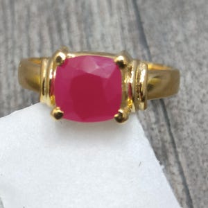 Op de afbeelding: Een goudkleurige ring met een grote, vierkante, robijnrode edelsteen. De ring heeft een klassiek ontwerp met decoratieve accenten op de band. De ring wordt gepresenteerd op een witte ondergrond.