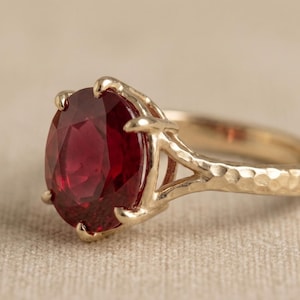 Peut inclure: Une bague en or avec une grande pierre précieuse rouge foncé de taille ovale maintenue par six griffes. L'anneau de la bague présente une finition martelée et texturée, et le sertissage rejoint l'anneau avec un design à double branche.
