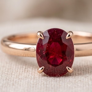 May include: A ring featuring a large, oval-cut, deep red ruby gemstone set in a polished rose gold band. The stone is secured by four prongs, highlighting the vibrant color and faceted surface of the gem against the smooth, metallic rose gold setting.