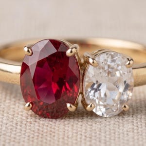 May include: A gold ring featuring two oval-cut stones set side-by-side. The larger stone is a deep red ruby, and the smaller stone is a clear, colourless diamond. Both stones are held in place by four-prong settings on a smooth, polished gold band.