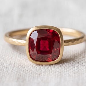 Pode incluir: Um anel de ouro com uma pedra preciosa vermelho-escura em corte almofada, engastada num aro liso. A banda do anel tem um acabamento martelado e texturizado, contrastando com o engaste polido da pedra central e a estrutura de ouro lisa.