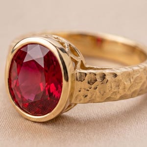 Pode incluir: Um anel de ouro com uma grande pedra preciosa de corte oval e cor vermelho-profundo, engastada num bisel liso. A banda do anel tem um acabamento martelado e texturizado, e a galeria sob a pedra central exibe um design de rede aberto.