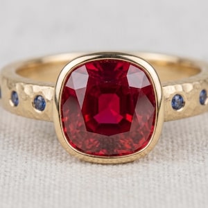 May include: A gold ring featuring a large, cushion-cut red gemstone set in a bezel. The band has a hammered texture and is accented with small, round blue gemstones embedded along the sides of the shank, creating a detailed, multi-stone design.