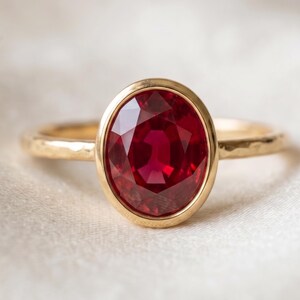 May include: A gold ring featuring a large, oval-cut, deep red ruby gemstone set in a smooth bezel. The ring band has a textured, hammered finish, providing a contrast to the polished gold setting that secures the vibrant, faceted red stone.