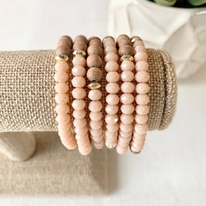 May include: A stack of six bracelets with light pink and wood beads. The bracelets are on a brown burlap surface.