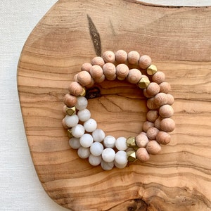May include: A set of three bracelets featuring wooden beads in a light brown color and white beads with gold accents. The bracelets are stacked on a wooden surface.