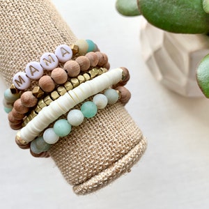 May include: A stack of bracelets with wooden, white, and green beads. The top bracelet has white beads with the word "MAMA" spelled out in gold letters.