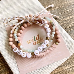 May include: Three beaded bracelets with the words "BESTIE" and "ELLE" spelled out in white letters on the beads. The bracelets are on a pink and white card with the text "wild oak essentials" and a website address. The bracelets are in a white drawstring bag.