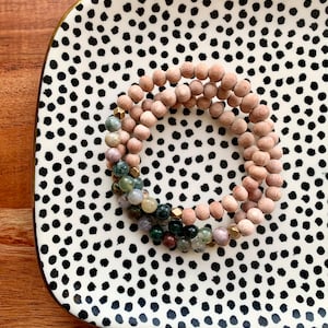 May include: A set of three bracelets on a black and white polka dot tray. The bracelets are made of wooden beads and gemstone beads in shades of green, brown, and purple.