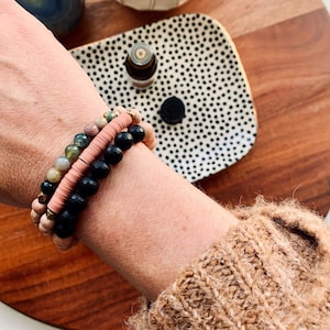 May include: A close-up of a person's wrist wearing three stacked bracelets. The bracelets are made of black beads, pink and green stones, and brown wood beads. The bracelets are accented with gold accents.