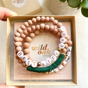 May include: A set of three wooden bead bracelets with the words "Best Life Ever" spelled out in white letters. The bracelets are in a small box with the brand name "Wild Oak Essentials" printed on the inside.