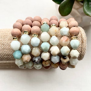 May include: A set of seven bracelets made with light green, brown, and white gemstone beads and wooden beads. The bracelets are stacked on a beige fabric surface.