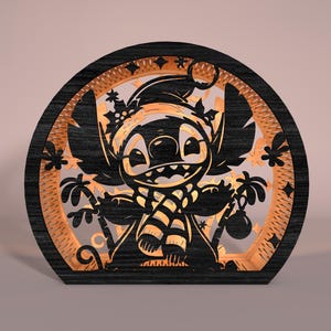 May include: A black and orange wooden laser-cut decoration featuring a cartoon character wearing a Santa hat and scarf. The design includes palm trees and ornaments, set within a circular frame. The character is smiling.