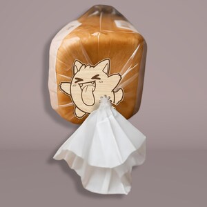 May include: A loaf of bread in a clear plastic bag, secured with a white twist tie. A wooden cat-shaped tag with a tongue sticking out is attached to the bag. The bread is golden brown.