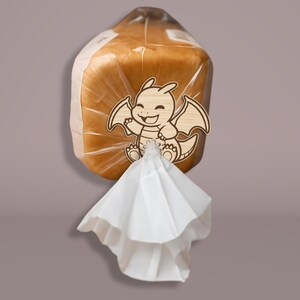 May include: A loaf of bread in a clear plastic bag, secured with a wooden dragon-shaped clip. The dragon has a smiling face and outstretched wings. A white paper bag is attached to the bottom of the bread.