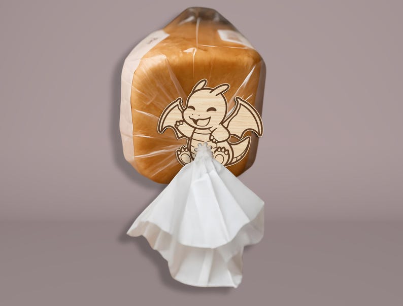 May include: A loaf of bread in a clear plastic bag, secured with a wooden dragon-shaped clip. The dragon has a smiling face and outstretched wings. A white paper bag is attached to the bottom of the bread.