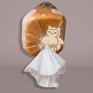 May include: A loaf of bread in a clear plastic bag, secured with a wooden bread clip. The clip is shaped like a cartoon character. A white paper napkin is attached to the clip.
