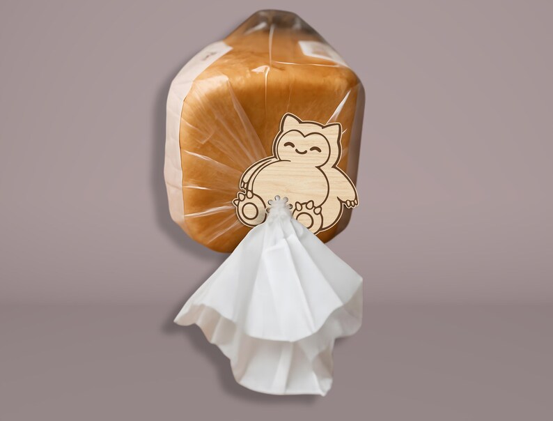 May include: A loaf of bread in a clear plastic bag, secured with a wooden bread clip. The clip is shaped like a cartoon character. A white paper napkin is attached to the clip.