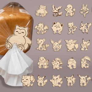 Puede incluir: Clips de bolsa de madera con temática de Pokémon y un clip para bolsa de pan. Los clips presentan varios personajes de Pokémon en color marrón claro. El clip para pan está sujeto a una bolsa de papel blanca. El fondo es gris liso.