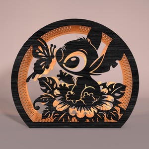 May include: A black and tan wooden decorative piece featuring a cartoon character with large eyes, a butterfly, and floral elements. The design is set within a circular frame with a textured inner border. The piece is likely a decorative item or a light fixture.