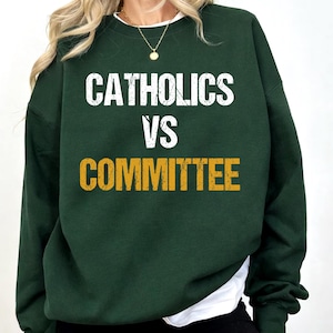 Op de afbeelding: Een donkergroene sweatshirt met de tekst "CATHOLICS VS COMMITTEE" in witte en gouden letters. De sweatshirt heeft lange mouwen en een ronde hals. Een wit shirt is eronder zichtbaar.