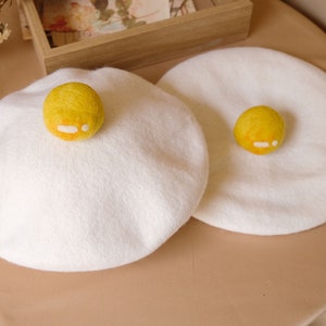 Op de afbeelding: Twee witte baretten met een geel dooierontwerp. De dooiers zijn gevilt en hebben een klein wit detail. De baretten zijn rond en liggen op een lichtbruin oppervlak. Een leuk en uniek accessoire.