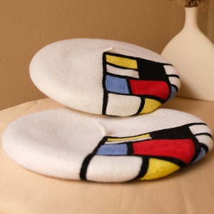 May include: Two white berets with a colorful geometric design. The design features blocks of red, yellow, blue, and black, outlined in black. One beret is stacked on top of the other. A white vase with dried flowers is in the background.
