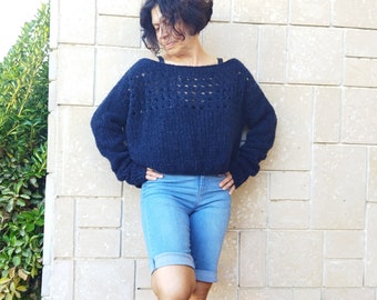 May include: A navy blue sweater with a wide neckline and open knit detailing across the chest. The sweater has long sleeves and is paired with light blue denim shorts. The person is leaning against a light-colored, square-tiled wall.