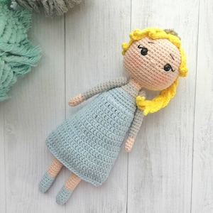 K&ouml;nnte beinhalten: Handgemachte H&auml;kelpuppe mit gelben Haaren, die ein hellblaues Kleid und ein graues Oberteil tr&auml;gt. Die Puppe hat ein s&uuml;&szlig;es Gesicht mit schwarzen Augen und rosigen Wangen. Die Arme und Beine der Puppe sind hellbeige. Die Puppe liegt auf einer wei&szlig;en Holzoberfl&auml;che.