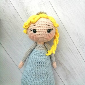 K&ouml;nnte beinhalten: Handgefertigte H&auml;kelpuppe mit hellem Hautton, gelben Haaren und blauem Kleid. Die Puppe hat eine kleine silberne Krone und rosige Wangen. Die Arme und der Oberk&ouml;rper sind grau. Ein bezaubernder handgefertigter Artikel.