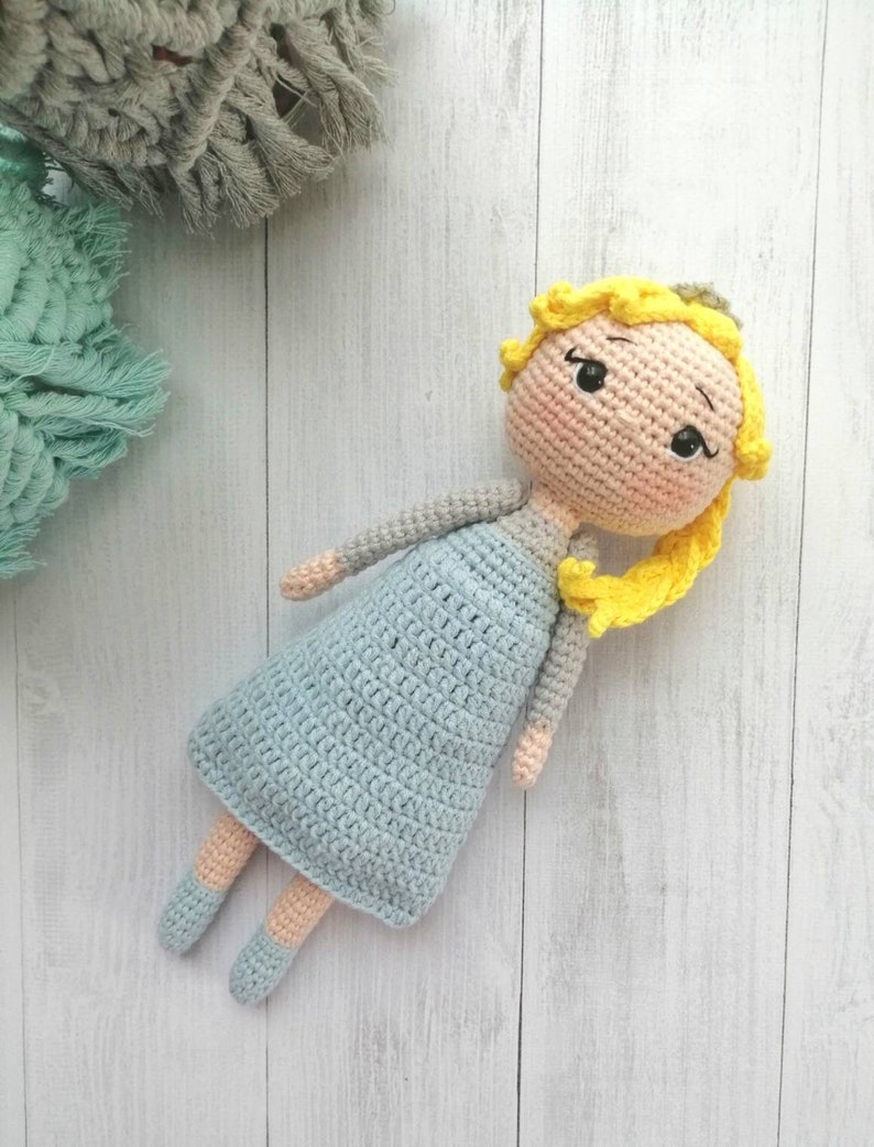 K&ouml;nnte beinhalten: Handgemachte H&auml;kelpuppe mit gelben Haaren, die ein hellblaues Kleid und ein graues Oberteil tr&auml;gt. Die Puppe hat ein s&uuml;&szlig;es Gesicht mit schwarzen Augen und rosigen Wangen. Die Arme und Beine der Puppe sind hellbeige. Die Puppe liegt auf einer wei&szlig;en Holzoberfl&auml;che.