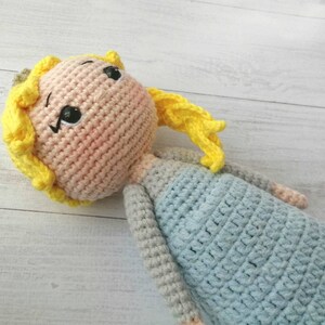 K&ouml;nnte beinhalten: Handgemachte H&auml;kelpuppe mit hellrosa Gesicht, gelben Haaren und einem hellblauen Kleid. Die Puppe hat schwarz gestickte Augen und einen kleinen, rosigen Rouge auf den Wangen. Die Arme und Beine der Puppe sind grau.
