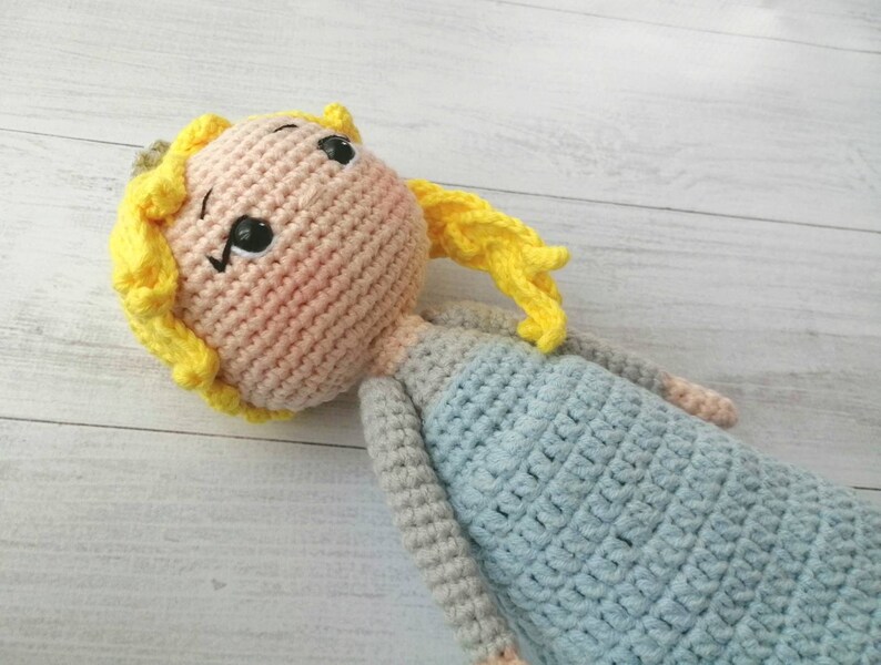K&ouml;nnte beinhalten: Handgemachte H&auml;kelpuppe mit hellrosa Gesicht, gelben Haaren und einem hellblauen Kleid. Die Puppe hat schwarz gestickte Augen und einen kleinen, rosigen Rouge auf den Wangen. Die Arme und Beine der Puppe sind grau.
