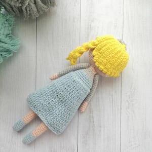 K&ouml;nnte beinhalten: Handgemachte H&auml;kelpuppe mit gelben Haaren, einem hellblauen Kleid und grauen Armen. Die Beine der Puppe sind hellblau und pfirsichfarben. Die Puppe liegt auf einer wei&szlig;en Holzoberfl&auml;che. Handgemachtes Spielzeug.