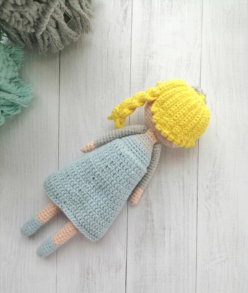 K&ouml;nnte beinhalten: Handgemachte H&auml;kelpuppe mit gelben Haaren, einem hellblauen Kleid und grauen Armen. Die Beine der Puppe sind hellblau und pfirsichfarben. Die Puppe liegt auf einer wei&szlig;en Holzoberfl&auml;che. Handgemachtes Spielzeug.