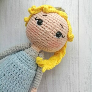 K&ouml;nnte beinhalten: Handgemachte H&auml;kelpuppe mit hellpfirsichfarbenem Gesicht, schwarzen Augen und gelben Haaren. Die Puppe tr&auml;gt ein hellblaues Kleid und hat graue Arme. Die Haare der Puppe sind zu einem seitlichen Pferdeschwanz gestylt. Handgemachtes Spielzeug.