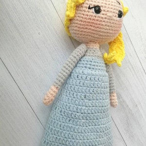 K&ouml;nnte beinhalten: Handgemachte H&auml;kelpuppe mit hellblauem Kleid, gelben Haaren und einem pfirsichfarbenen Gesicht und Gliedma&szlig;en. Die Puppe hat schwarze Augen und ist vor einem hellen Holzmaserungshintergrund drapiert. Ein handgefertigtes Spielzeug.