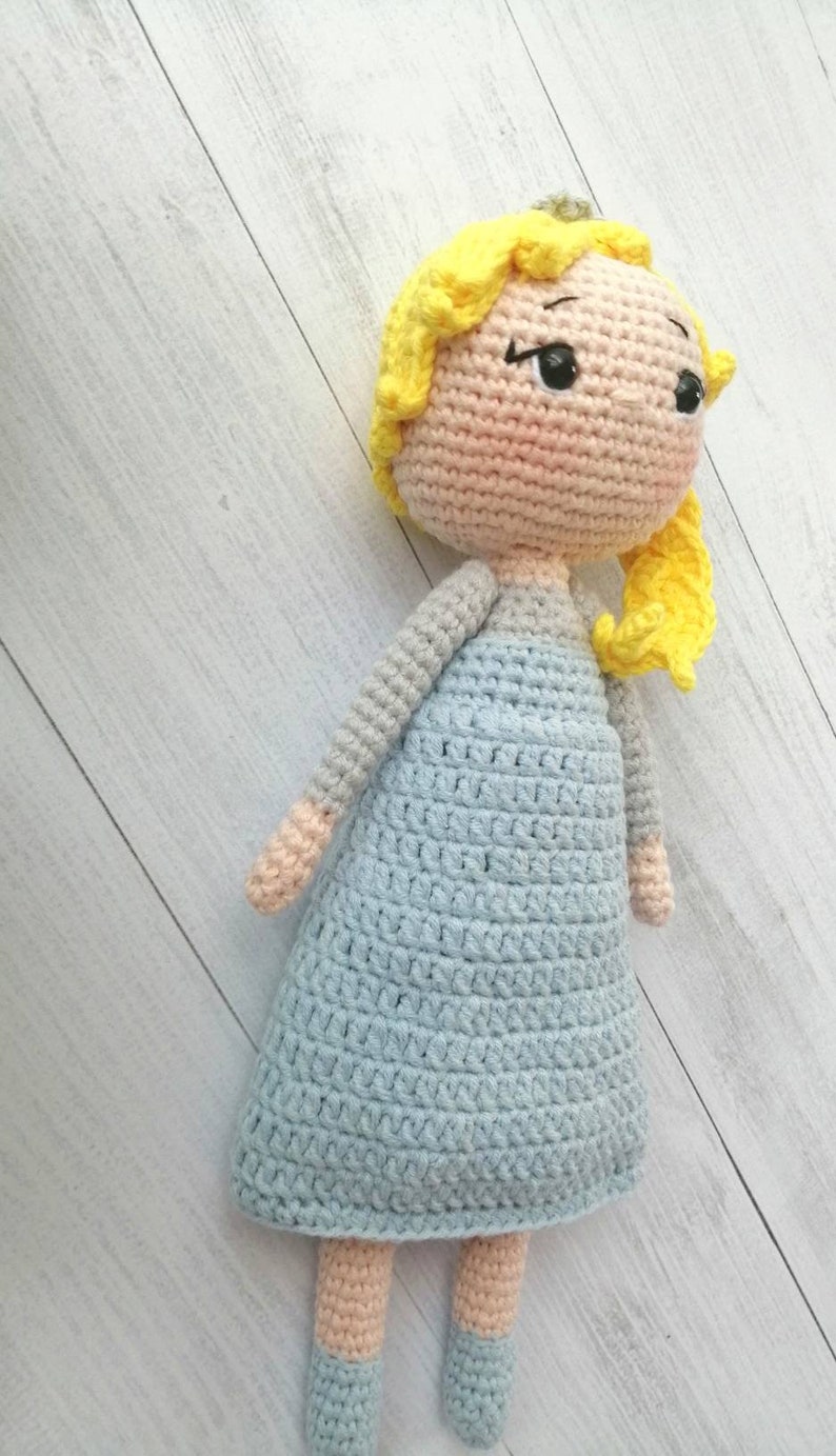K&ouml;nnte beinhalten: Handgemachte H&auml;kelpuppe mit hellblauem Kleid, gelben Haaren und einem pfirsichfarbenen Gesicht und Gliedma&szlig;en. Die Puppe hat schwarze Augen und ist vor einem hellen Holzmaserungshintergrund drapiert. Ein handgefertigtes Spielzeug.