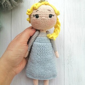 K&ouml;nnte beinhalten: Handgefertigte H&auml;kelpuppe mit hellem Hautton, gelben Haaren und einem hellblauen Kleid. Die Puppe hat schwarze Augen und tr&auml;gt ein hellblaues Kleid. Die Puppe ist etwa 20 cm gro&szlig;.