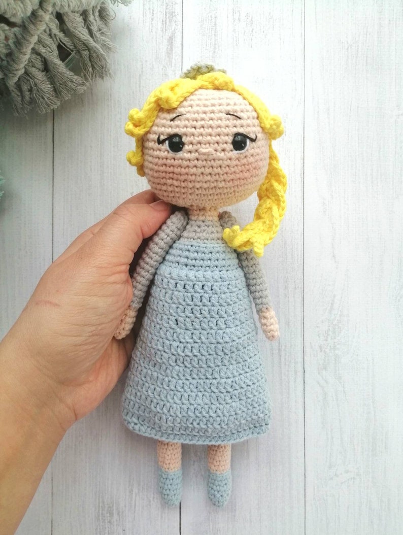 K&ouml;nnte beinhalten: Handgefertigte H&auml;kelpuppe mit hellem Hautton, gelben Haaren und einem hellblauen Kleid. Die Puppe hat schwarze Augen und tr&auml;gt ein hellblaues Kleid. Die Puppe ist etwa 20 cm gro&szlig;.